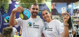 A man and a woman in Fairtrade t-shirts holding up a Fairtrade logo sticker and a cup, at a coffee trade show.